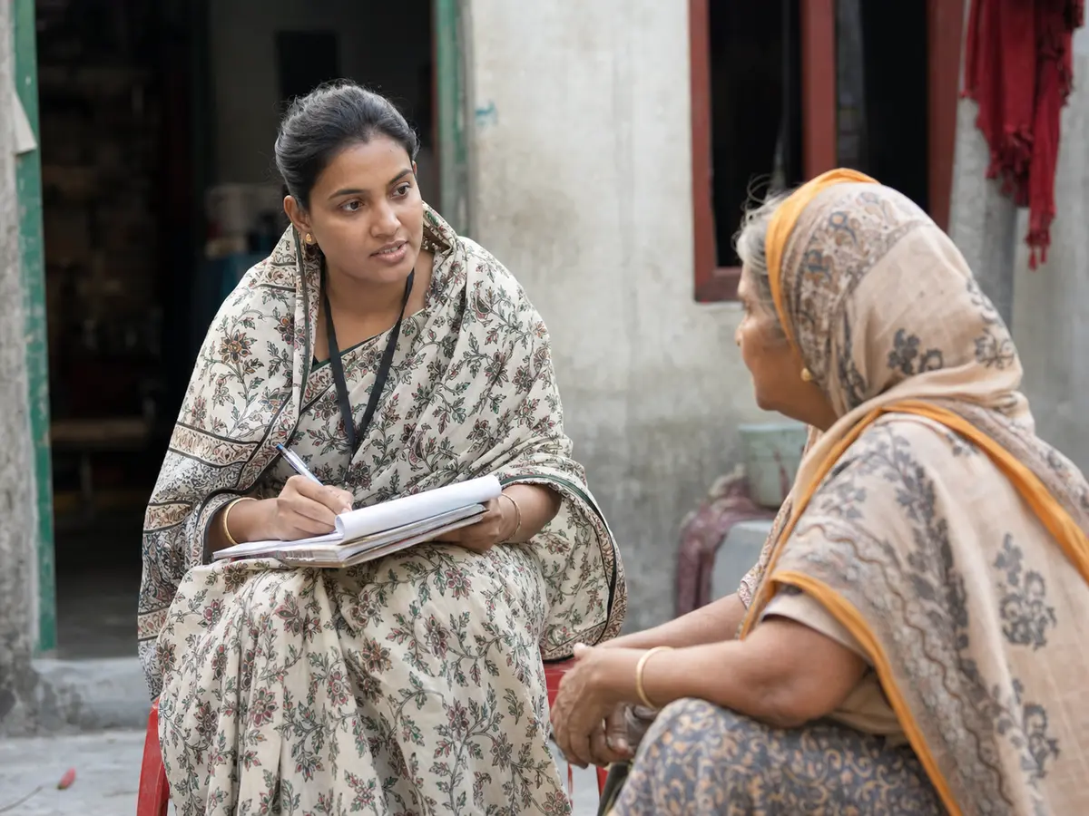 A community health worker takes notes during a courtyard conversation.